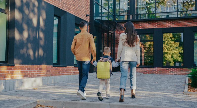 Young family spending time together while walking outdoors near contemporary building with red brick walls and black window frames. Child going between parents. Ready for school with his backpack.